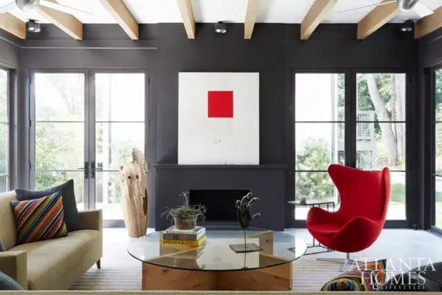Living room with dark gray walls, exposed wooden beams, a black fireplace with abstract art, a red egg chair, and a glass coffee table with books and a sculpture.