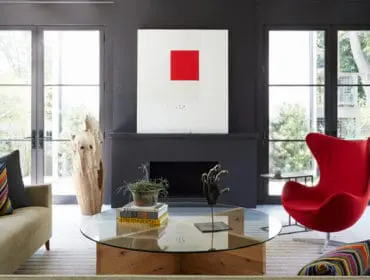 Living room with a tan sofa, a red egg chair, a glass coffee table with books and a sculpture, and a dark gray fireplace with abstract art above it.