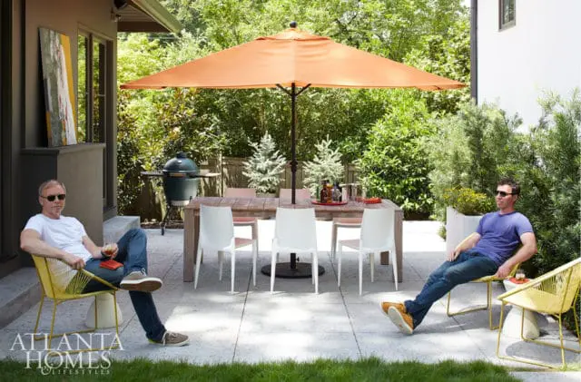 Two men relax on a patio with a large orange umbrella, dining table, white chairs, and a green grill.