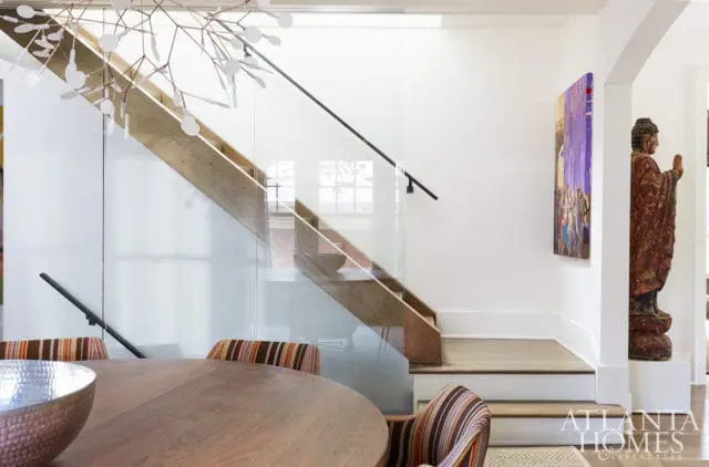 Modern staircase with glass railing and wood treads, next to a dining table with striped chairs and a large wooden Buddha statue.