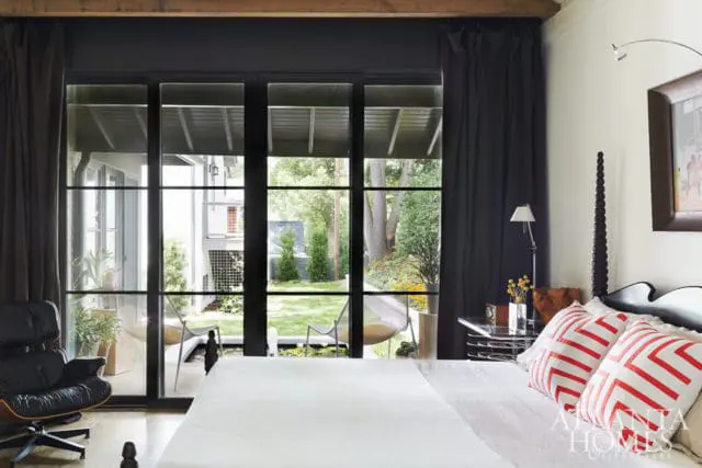 Bedroom with black Eames chair, large black-framed windows overlooking a garden, and a bed with white linens and red-and-white striped pillows.