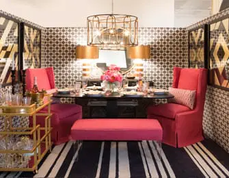 Dining room with oval table set for four, flanked by two coral upholstered wingback chairs and a coral bench. Geometric wallpaper, striped rug, and gold bar cart.