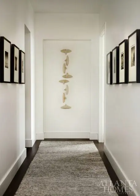 Hallway with dark wood floors, a grey textured runner rug, and white walls adorned with framed black and white photographs and a vertical wooden sculpture.