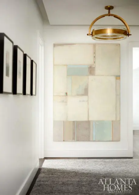 Hallway with abstract art, framed pictures, and a brass chandelier. The art features a mosaic of cream, beige, and pale blue rectangles.