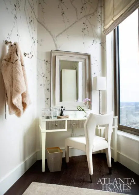 Corner vanity with a light-colored upholstered chair, a mirror, and a faux fur coat hanging on the wall with floral wallpaper.