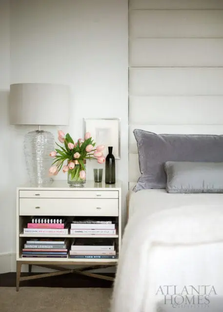 Bedroom with a white upholstered headboard, grey pillows, and a white nightstand holding a glass lamp, pink tulips, and books.