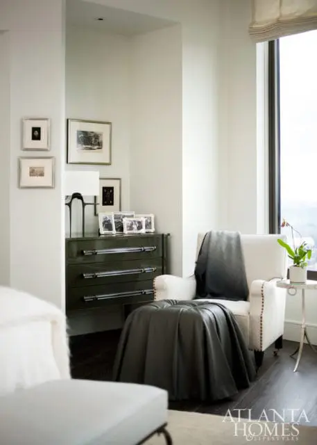 White armchair with a dark gradient throw blanket draped over it, next to a dark green dresser with clear drawer pulls. Framed artwork hangs on the wall above the dresser.