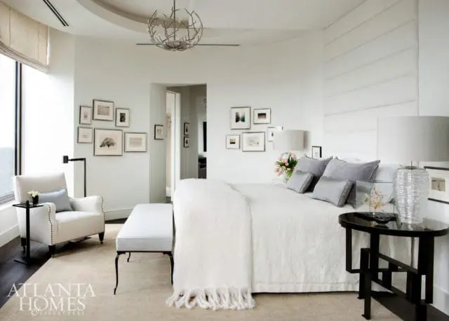Serene bedroom with a large white bed, grey pillows, and a white textured headboard. A white armchair and bench sit on a neutral rug. Framed art adorns the walls, and a branch-like chandelier hangs from the ceiling.