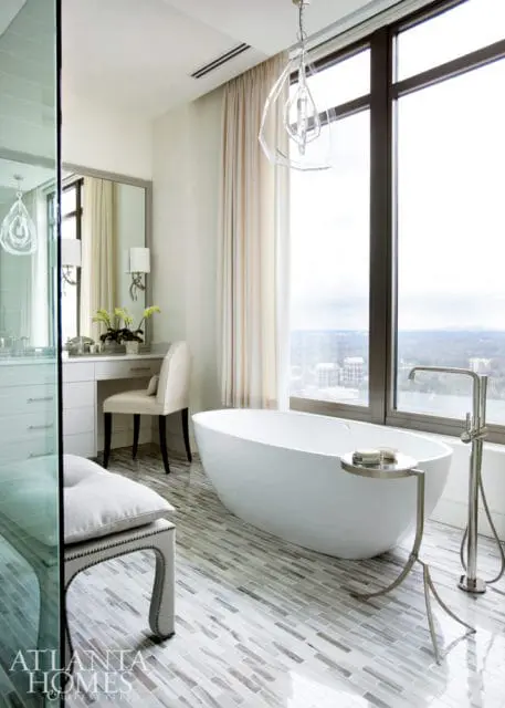 Modern bathroom with freestanding white bathtub, vanity with mirror, and mosaic tile flooring. Large window overlooks city skyline.