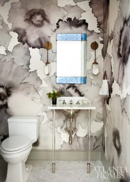 Powder room with large-scale floral wallpaper in shades of gray and white, a white toilet, a pedestal sink with chrome fixtures, and a mirror with a blue decorative frame.