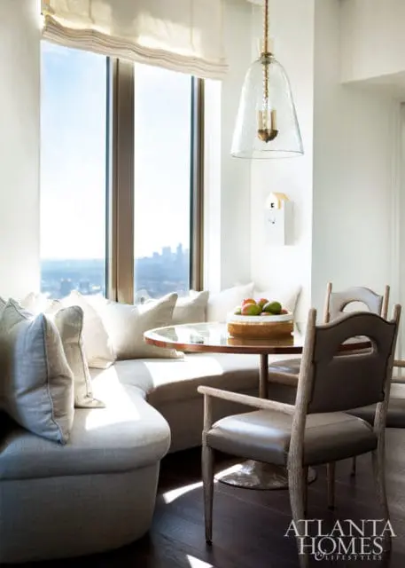 Bright breakfast nook with curved built-in sofa, round wood table, and leather chairs, featuring a large window with city view and a glass pendant light.