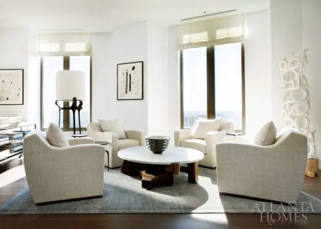 Living room with four cream upholstered armchairs arranged around a round marble coffee table with a dark wood base. Large windows with sheer blinds overlook a city skyline.