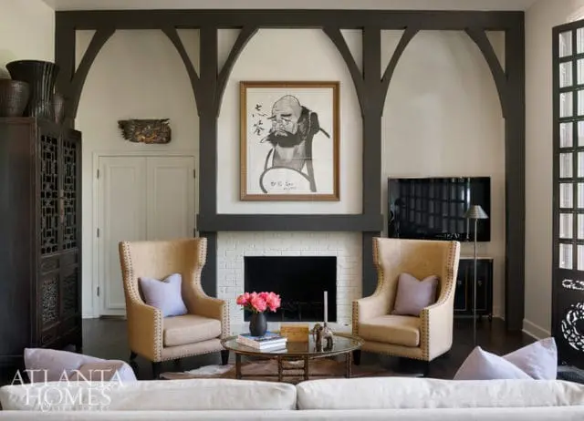Living room with a white brick fireplace, a framed painting of a man above the mantel, two upholstered wingback chairs, and a dark wood cabinet.