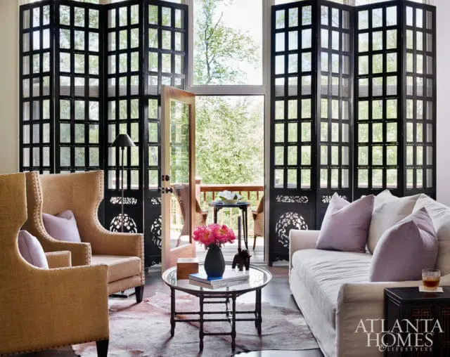 Living room with a large black folding screen, tan wingback chairs, a white sofa with lavender pillows, and a glass coffee table with flowers.