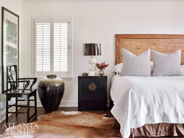 Bedroom with white walls, white plantation shutters, a dark wood bedside table with a lamp and flowers, a large dark ceramic vase, a black chair, and a bed with a wooden headboard and striped pillows.