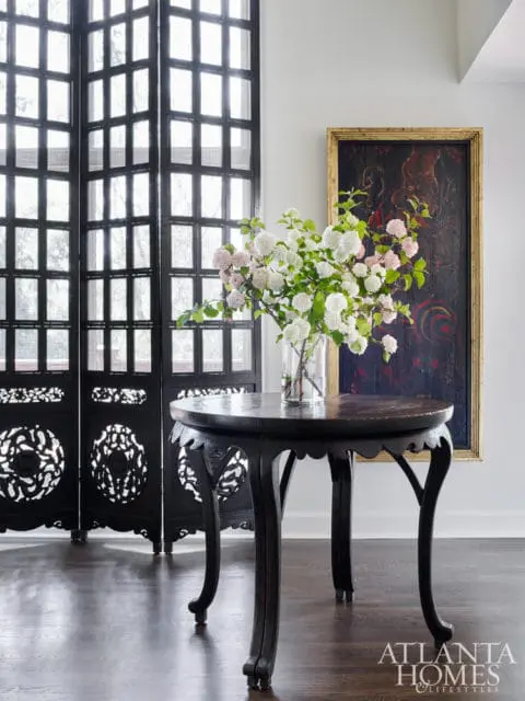 Dark wood round table with carved legs holding a vase of white and pink flowers, next to a black folding screen and a framed painting.