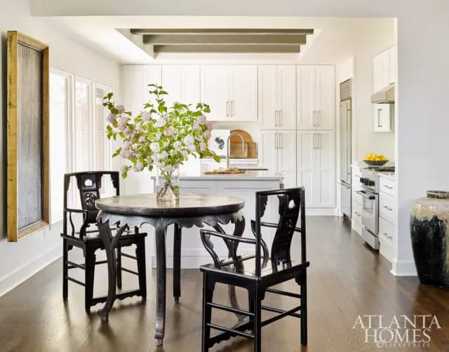 Dining area with antique chairs and table in front of a white kitchen with stainless steel appliances and a large vase.