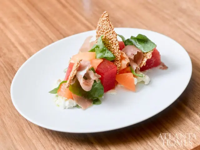 White plate with a salad of cubed watermelon, cantaloupe, prosciutto, arugula, and a sesame seed cracker on a wooden table.