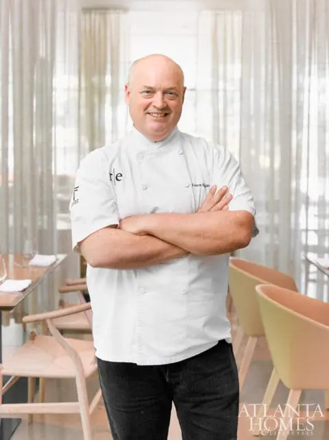 Chef Linton Hopkins smiles with arms crossed in a restaurant dining room, wearing a white chef's coat and dark jeans.