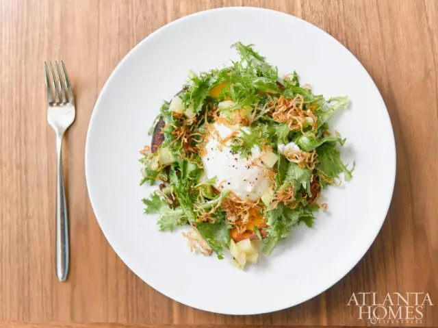 White plate with a salad topped with a poached egg and crispy fried onions, next to a silver fork on a wooden table. Atlanta Homes & Lifestyles logo in the bottom right corner.