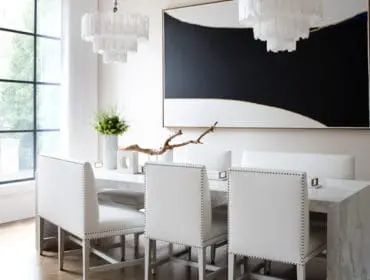 Modern dining room with white upholstered chairs around a marble table, featuring a large abstract black and white painting and tiered chandeliers.