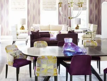 Dining room with oval dark wood table and purple velvet chairs, featuring patterned wallpaper, a crystal chandelier, and a living area with sofas and accent chairs.