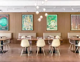 Modern dining room with wood-paneled walls, built-in banquettes, and colorful abstract art. Cream-colored chairs surround wooden tables under a cluster of globe pendant lights.