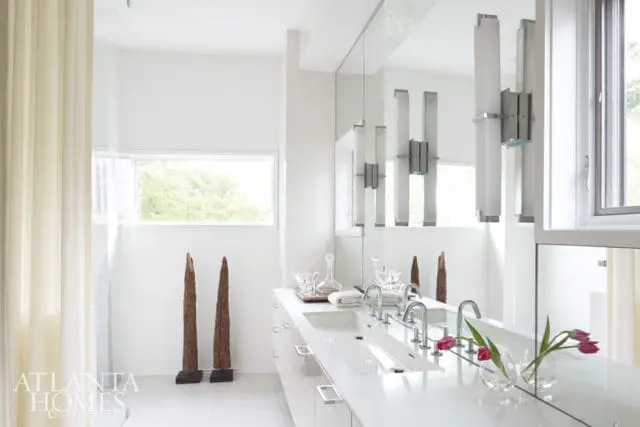 Modern white bathroom with a long double vanity, chrome faucets, and a large mirror reflecting vertical sconces. Two tall wooden sculptures stand near a window.