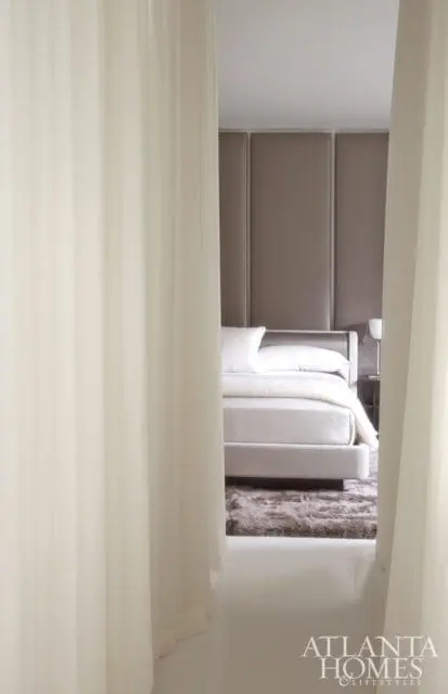 Bedroom with a plush gray rug, a light gray upholstered bed with white linens, and a taupe upholstered headboard wall. Sheer white curtains frame the view.