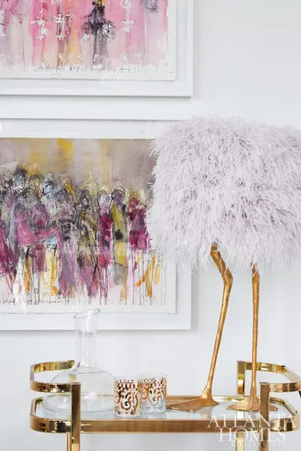Gold bar cart with glass shelves holding a decanter and two gold-patterned glasses, topped with a fluffy pink ostrich lamp and framed abstract art on the wall.