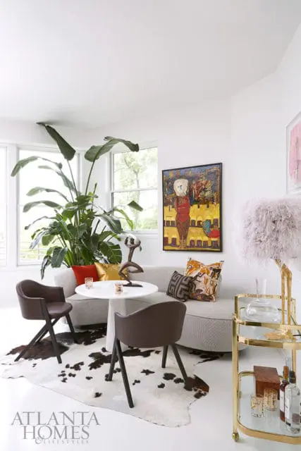 Modern living area with a curved gray sofa, white pedestal table, two brown chairs on a cowhide rug, and a large potted plant. Artwork hangs on the wall above the sofa.