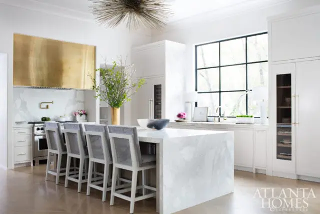 Modern white kitchen with a large marble island, brass range hood, gray upholstered bar stools, and a large black-framed window.