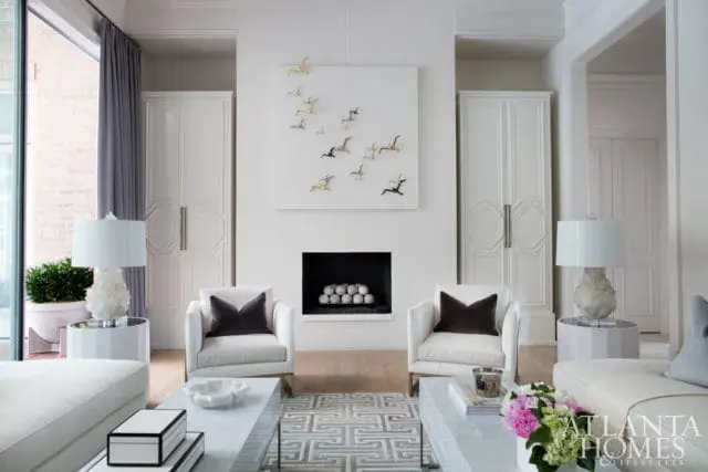 White living room with a fireplace featuring a white square art piece with gold and black bird sculptures, flanked by tall white cabinets. Two white armchairs with dark pillows face the fireplace.