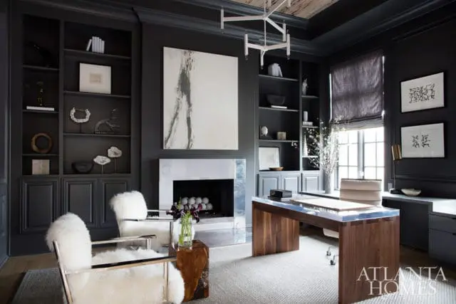 Dark gray office with built-in bookshelves, a modern fireplace with a silver surround, a large abstract painting, a wooden desk with a glass top, and two white sheepskin chairs.