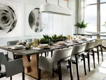 Modern dining room with a long concrete table set for a meal, surrounded by upholstered chairs. Large abstract art hangs on the wall above a planter filled with greenery.