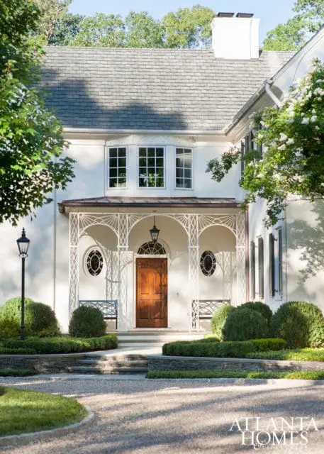 White stucco house with a gray slate roof, a wooden front door, and a decorative white porch with benches. Landscaped garden with gravel driveway and stone steps.