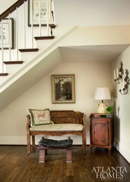Wooden bench with upholstered seat and decorative pillow beneath a staircase landing, next to a small wooden cabinet with a lamp and books.