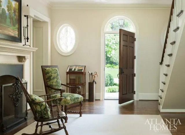 Foyer with a fireplace, two chairs upholstered in green botanical print fabric, an open wooden door revealing a garden, and a staircase.