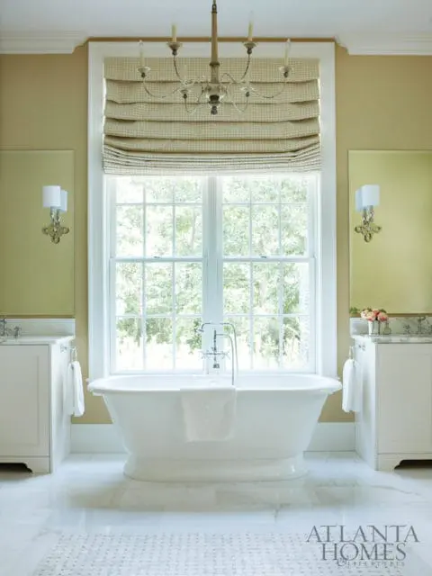 White freestanding bathtub in a bathroom with marble floors, double vanities, and a large window overlooking trees.