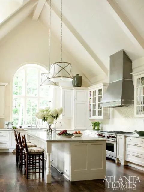 White kitchen with vaulted ceiling, large arched window, marble island with bar stools, and stainless steel range hood.