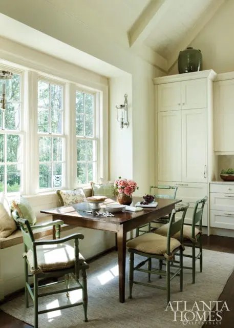 Dining nook with a wooden table, green chairs, and a built-in banquette under large windows. Cream cabinetry and a vaulted ceiling are visible.