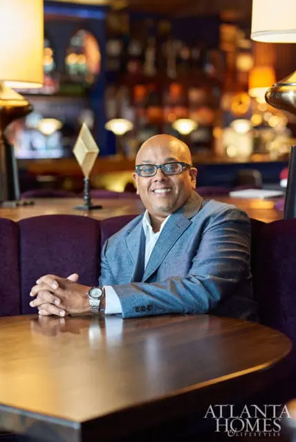 Man in a grey suit and glasses sitting at a table in a restaurant with purple upholstery and warm lighting.