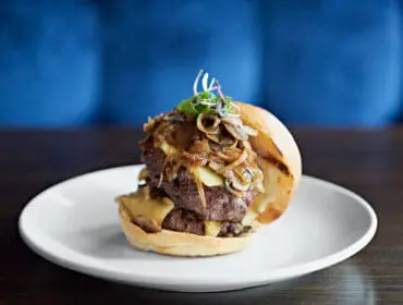 Double cheeseburger with sauteed mushrooms and onions on a white plate with a blurred blue background.
