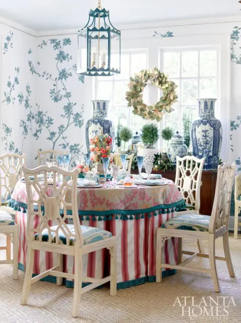 Dining room with blue floral wallpaper, a table set for a meal with a striped skirt and fringed tablecloth, and chinoiserie-style chairs.