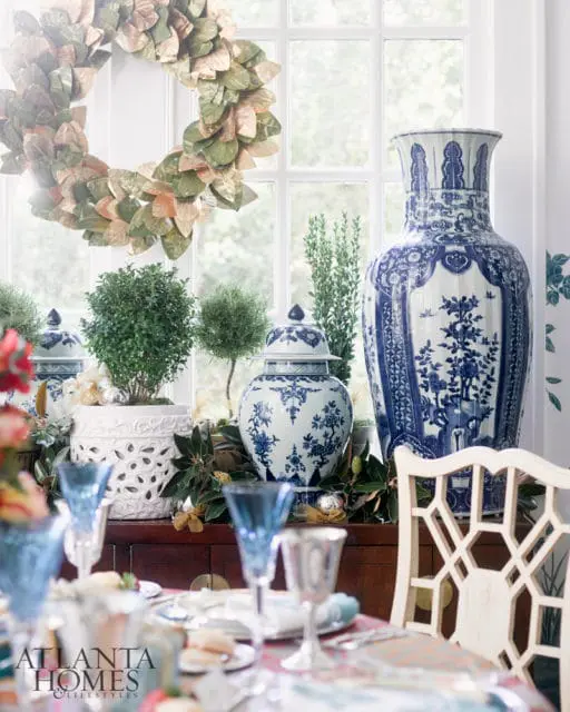 Dining room table set with blue glassware and silver goblets, featuring a large blue and white ginger jar and a decorative wreath on a window