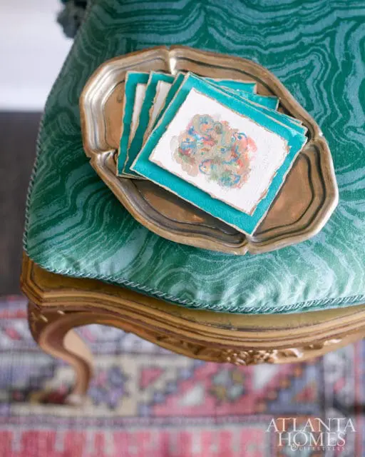 Stack of small watercolor paintings with torn paper edges on a tarnished silver tray, resting on a green patterned upholstered chair with gold trim.