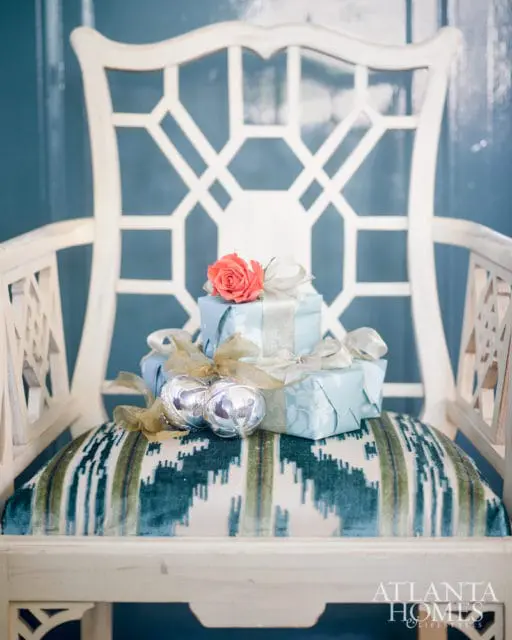 White lattice-back chair with a patterned seat cushion holding two light blue wrapped gifts topped with a coral rose and silver ornaments.