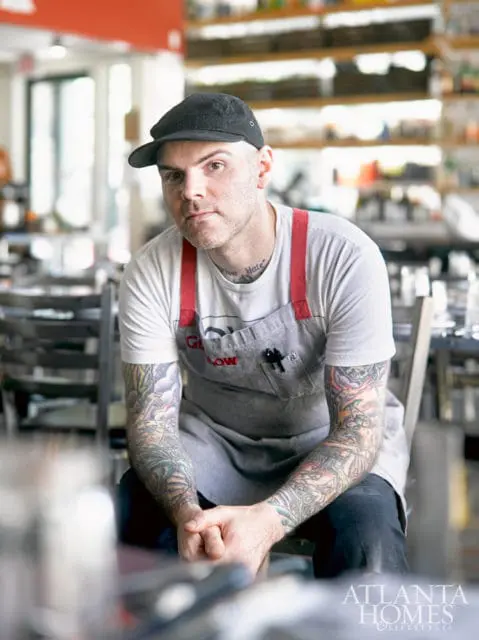 Man with tattoos on his arms and neck, wearing a black cap, white t-shirt, and grey apron, sitting in a restaurant setting.