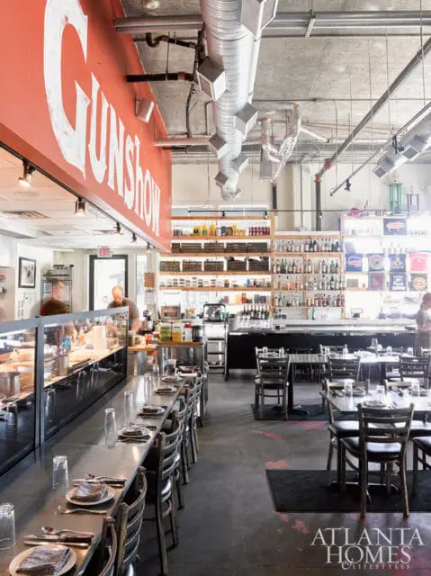 Restaurant interior with a long metal bar, tables set for dining, and shelves stocked with bottles and jars. A red sign with white text reads "GUNSHOW."