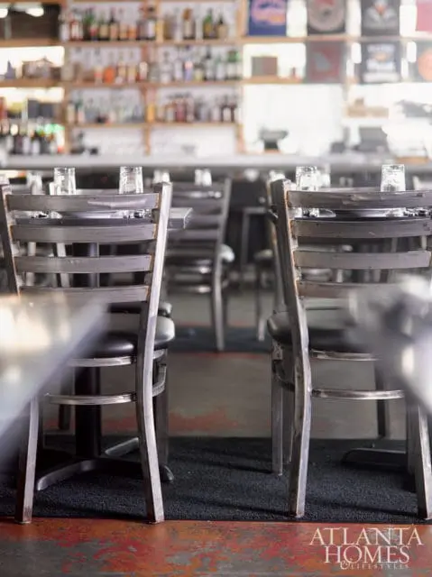 Restaurant interior with dark metal chairs and tables set with glasses, blurred shelves of bottles in the background, and a logo in the corner.